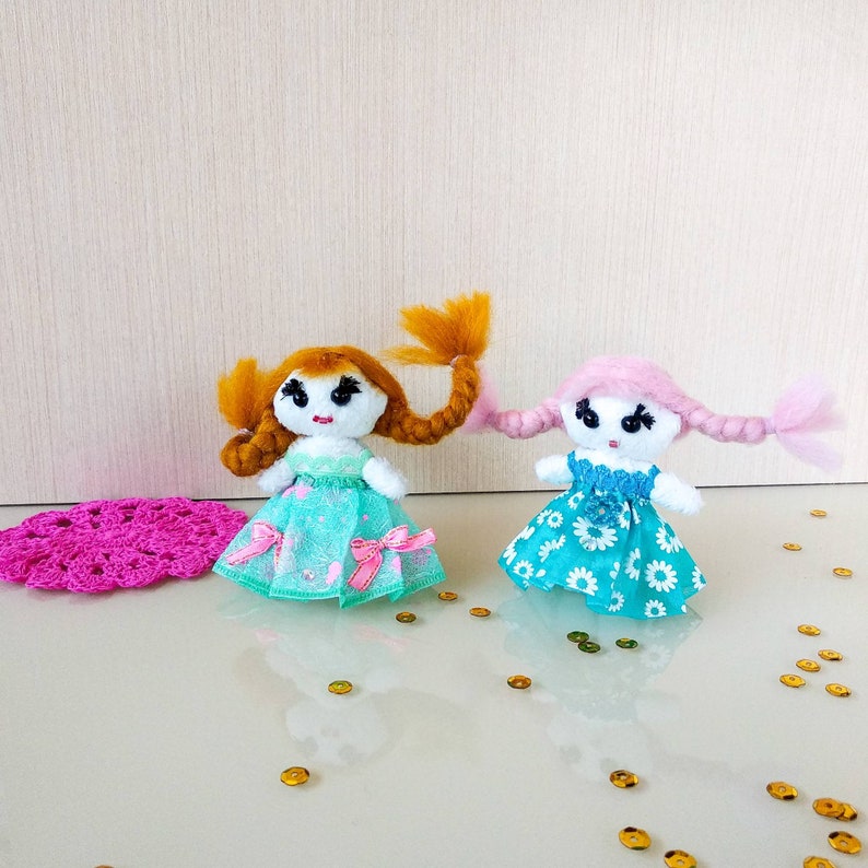 Op de afbeelding: Twee handgemaakte poppen met garen haar, gekleed in jurken. E&eacute;n pop heeft oranje haar en een turquoise jurk met roze strikken. De andere pop heeft roze haar en een blauwe bloemenjurk. Een roze gehaakte mat en gouden pailletten zijn ook in de afbeelding.