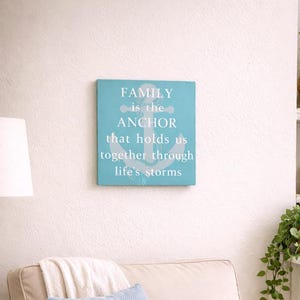 Coastal wood sign with painted in the Gulf with gray anchor design and quote “family is the anchor that holds us together through life’s storms,” displayed above a cozy armchair in a beach-style living room nook with soft neutral decor.