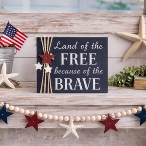 Rustic farmhouse mantel styled with patriotic décor, featuring a navy wood sign that reads “Land of the Free because of the Brave,” accented with twine and star embellishments, surrounded by American flags, a red lantern, greenery, and wooden bead garland.
