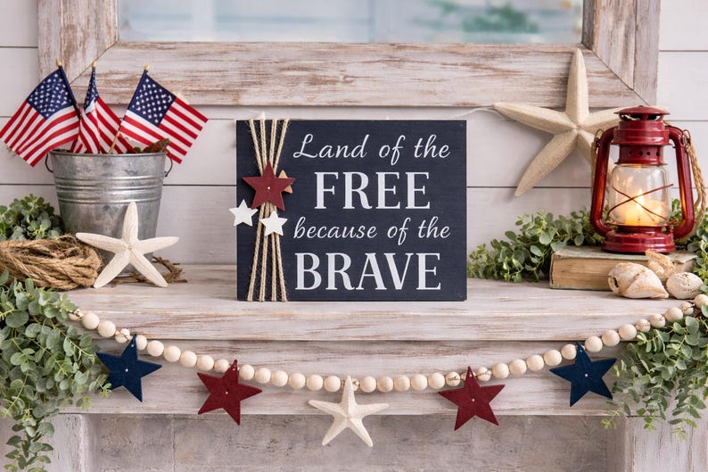 Rustic farmhouse mantel styled with patriotic décor, featuring a navy wood sign that reads “Land of the Free because of the Brave,” accented with twine and star embellishments, surrounded by American flags, a red lantern, greenery, and wooden bead garland.