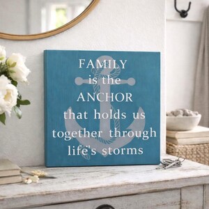 Turquoise coastal wood sign with gray anchor and white lettering reading “family is the anchor that holds us together through life’s storms,” displayed on a distressed white entryway table with flowers, books, and decorative accents.