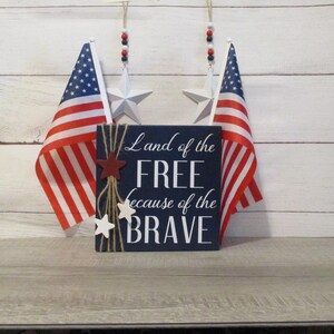 A blue wooden sign with white text that reads "Land of the FREE because of the BRAVE". The sign is decorated with red and white stars and twine. Two American flags are hanging on the wall behind the sign.