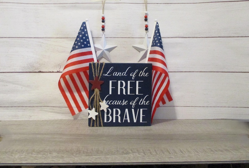 A blue wooden sign with white text that reads "Land of the FREE because of the BRAVE". The sign is decorated with red and white stars and twine. Two American flags are hanging on the wall behind the sign.