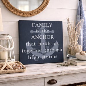 Navy blue coastal wood sign with anchor design and quote “family is the anchor that holds us together through life’s storms,” displayed on a rustic entryway table with nautical decor including rope lantern, seashells, driftwood, and woven accents.