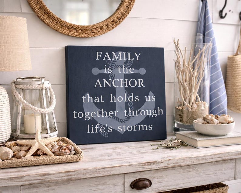 Navy blue coastal wood sign with anchor design and quote “family is the anchor that holds us together through life’s storms,” displayed on a rustic entryway table with nautical decor including rope lantern, seashells, driftwood, and woven accents.