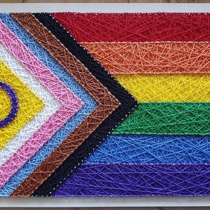Könnte beinhalten: Ein String-Art-Stück mit einem farbenfrohen Design mit einem Regenbogen aus Farben, darunter Rot, Orange, Gelb, Grün, Blau und Lila. Das Design enthält auch die Farben und Formen der Progress Pride Flagge.