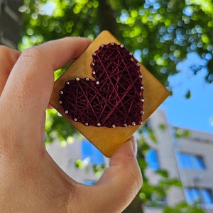 Könnte beinhalten: Ein handgefertigtes String-Art-Stück mit einem weinroten Herzdesign auf einer quadratischen Holzbasis. Das Herz wird mit weinrotem Faden um kleine silberne Stifte gewickelt. Die Holzbasis hat eine natürliche Holzmaserung.