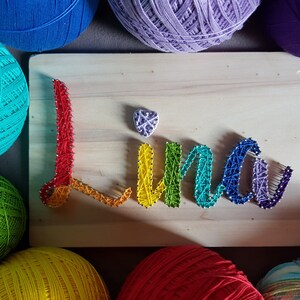 Könnte beinhalten: Ein Holzschild mit dem Namen "Lina" in farbenfroher String-Art. Die Buchstaben sind in Regenbogenfarben gehalten, mit einem kleinen Herz über dem "i". Garnknäuel in verschiedenen Farben umrahmen das Schild.