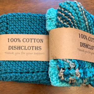 May include: Two knitted cotton dishcloths, one teal blue and one with a teal blue, light blue, and gray pattern. Both have a brown tag that reads "100% Cotton Dishcloths Thank you for your support!"