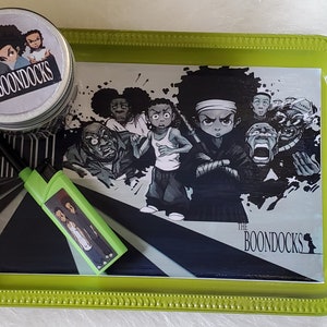 K&ouml;nnte beinhalten: Ein gr&uuml;nes Tablett mit einer Cartoon-Grafik von Charakteren aus The Boondocks. Ein passendes Feuerzeug und eine kleine Dose sind ebenfalls vorhanden. Das Tablett, das Feuerzeug und die Dose zeigen das Logo und die Charakterkunst der Show.