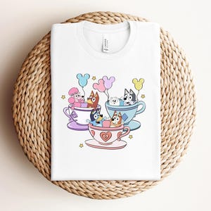 Op de afbeelding: Wit T-shirt met een kleurrijke cartoonillustratie van honden in theekopjes, met ballonnen. Het ontwerp bevat een poedel, een husky en andere hondenrassen. De theekopjes zijn in pastelkleuren met hart- en steraccenten.