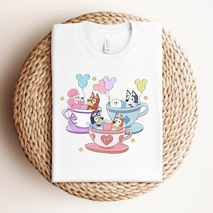 以下が含まれることがあります： 白いTシャツに、ティーカップに座っている4匹の犬の漫画の画像が描かれています。犬たちはカラフルな帽子をかぶり、風船を持っています。ティーカップはハートと花で飾られています。