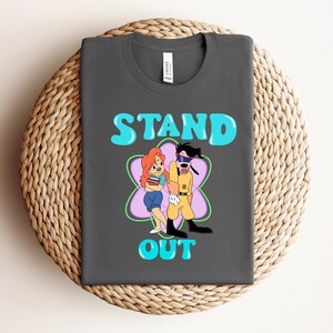 Può includere: Una maglietta grigio scuro con un'immagine di un cartone animato di un cane e una ragazza in piedi in un fiore viola. Il testo "STAND OUT" è stampato in blu sulla maglietta.