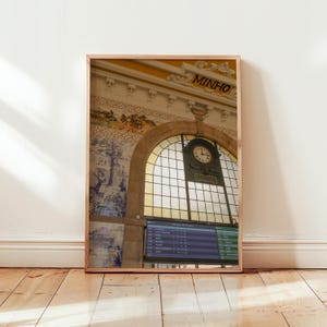 Puede incluir: Fotografía enmarcada de un interior de estación de tren. La imagen presenta una gran ventana arqueada con un reloj, un panel de salidas y azulejos decorativos. La palabra "MINHO" es visible encima de la ventana.