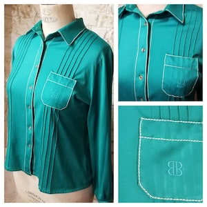 Puede incluir: Una camisa de manga larga de color azul verdoso con costuras blancas y un bolsillo en el lado izquierdo. El bolsillo tiene las letras "BB" bordadas.
