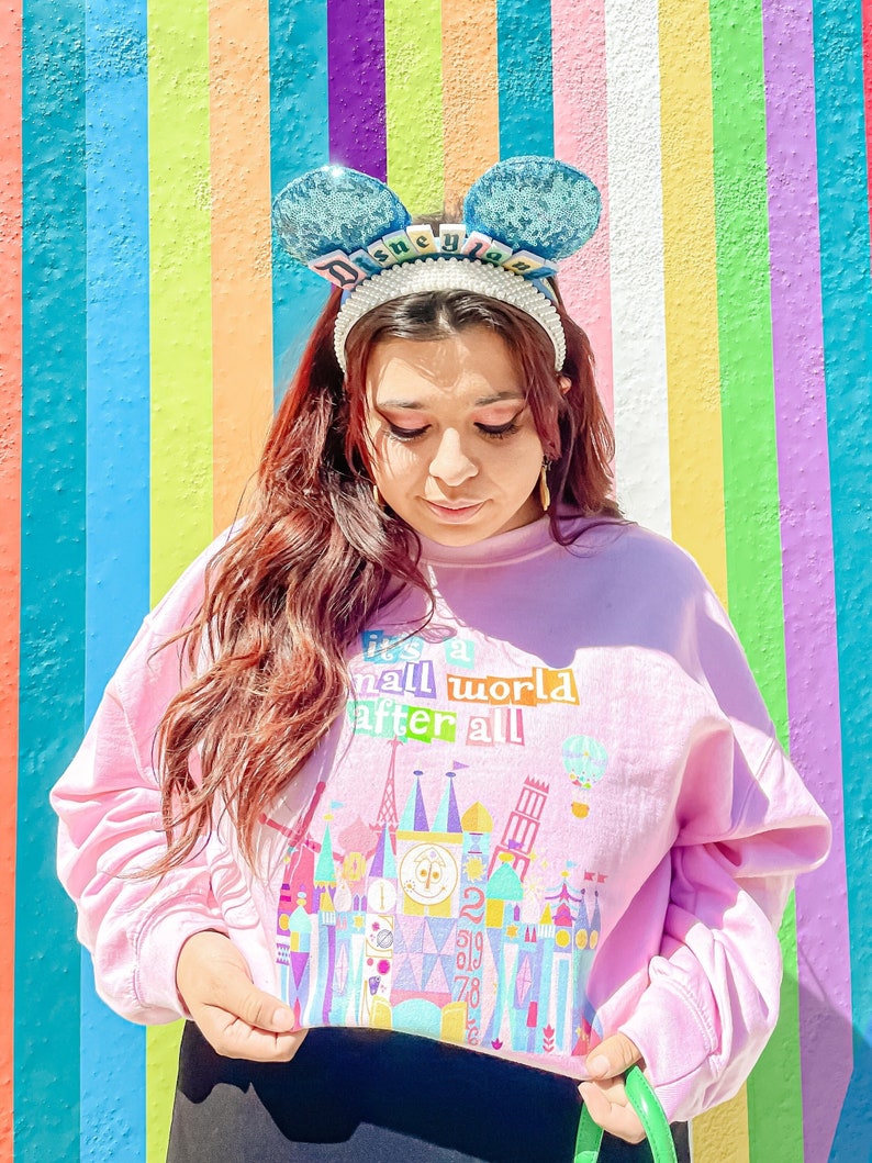 May include: A pink sweatshirt featuring a colourful graphic of the iconic Cinderella's Castle at Walt Disney World. The text "It's a small world after all" is printed above the castle. The sweatshirt has a cropped fit and is being worn by a person with blue and white Mickey Mouse ears.