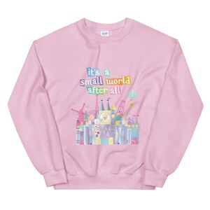 May include: Pink sweatshirt featuring a colourful graphic of the 'it's a small world' ride at Disney World. The graphic includes the text 'it's a small world after all'.