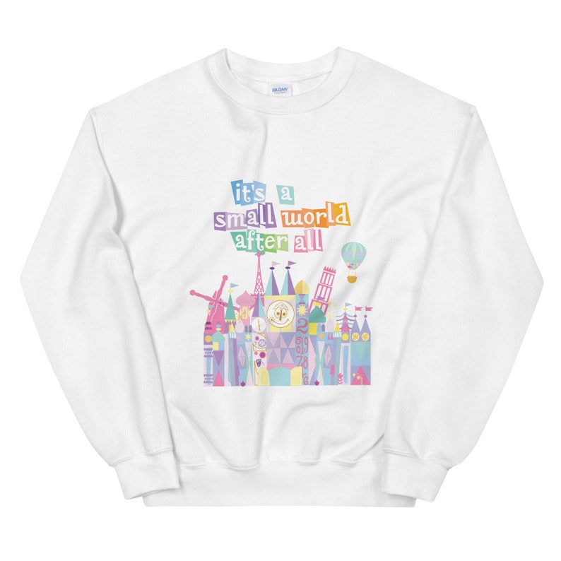 May include: White sweatshirt featuring a colourful graphic of the 'it's a small world' ride at Disney World. The graphic includes the ride's name in rainbow letters and a colourful illustration of the ride's facade.