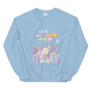 May include: Light blue sweatshirt featuring a colourful graphic of the "it's a small world" ride at Disney World. The graphic includes the text "it's a small world after all".