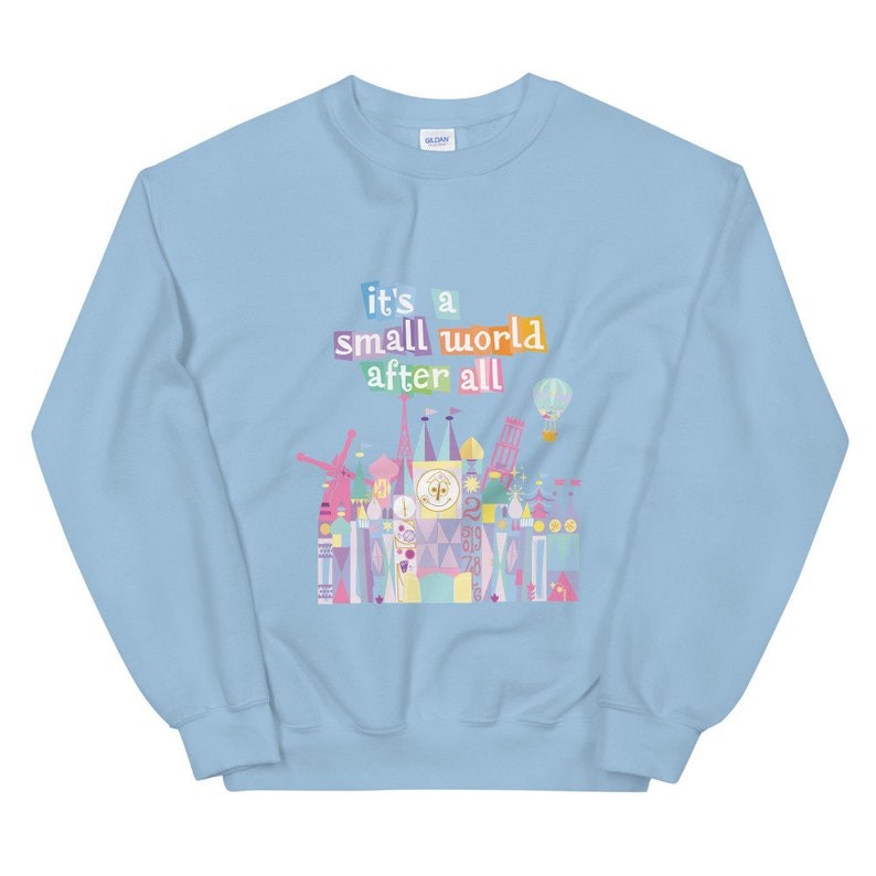May include: Light blue sweatshirt featuring a colourful graphic of the "it's a small world" ride at Disney World. The graphic includes the text "it's a small world after all".