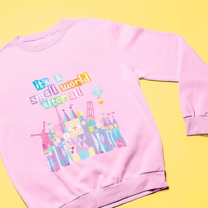 May include: Pink sweatshirt featuring a colourful graphic of the "it's a small world" ride at Disney World. The graphic includes the text "it's a small world after all".