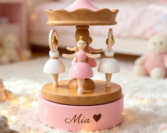 May include: A wooden carousel music box featuring three ballerina figures with white and pink dresses. The base is painted light pink and displays the name Mia followed by a heart symbol. The structure has a natural wood finish on the top canopy and central pillar, with a smooth, circular wooden platform supporting the rotating dancers.