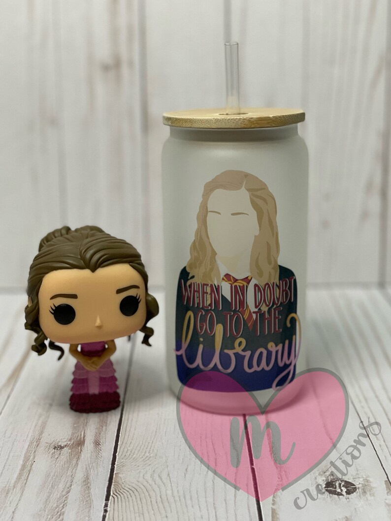 May include: A white frosted tumbler with a wooden lid and a straw. The tumbler has a graphic of a woman with the text "When in doubt go to the library".