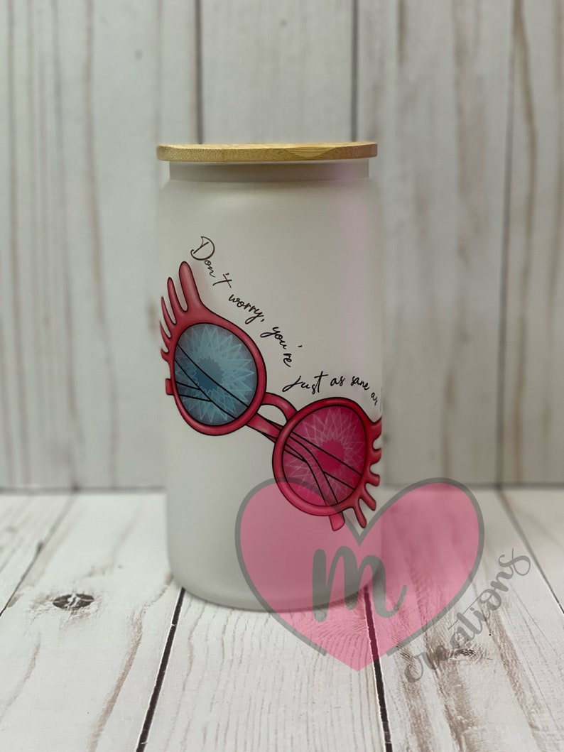 May include: White frosted glass jar with a wooden lid. The jar has a pink heart with the letter "m" inside and a graphic of pink glasses with blue lenses. The text on the jar reads "Don't worry, you're just as sane as me."