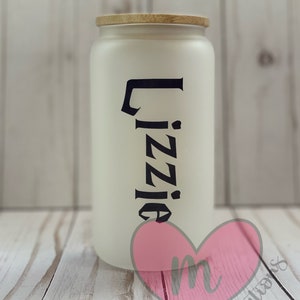 May include: White ceramic jar with a wooden lid and the name "Lizzie" written in black lettering. The jar is sitting on a wooden surface.