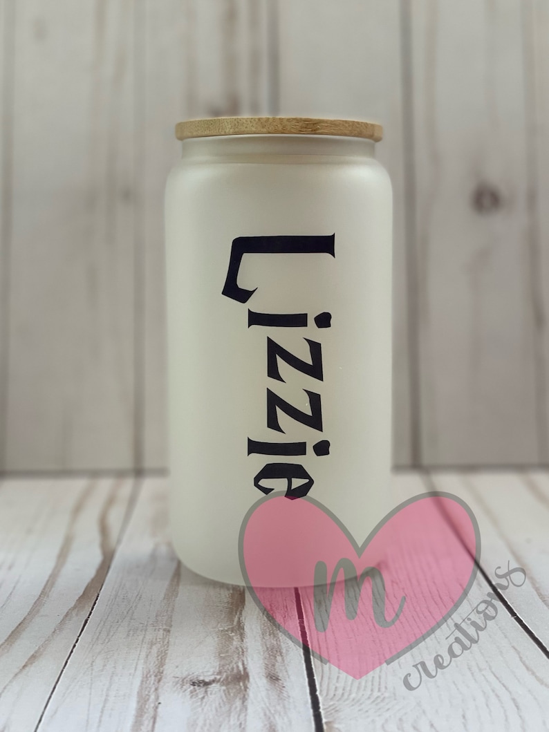 May include: White ceramic jar with a wooden lid and the name "Lizzie" written in black lettering. The jar is sitting on a wooden surface.