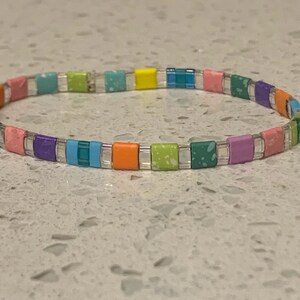 May include: A colorful beaded bracelet with a rainbow pattern. The bracelet is made of small, square beads in shades of pink, purple, blue, green, yellow, and orange.