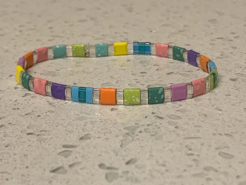 May include: A colorful beaded bracelet with a rainbow pattern. The bracelet is made of small, square beads in shades of pink, purple, blue, green, yellow, and orange.