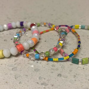 May include: Four colorful beaded bracelets, one with white beads, one with clear crystal beads, one with small square beads in a rainbow pattern, and one with a mix of blue, green, and orange beads.