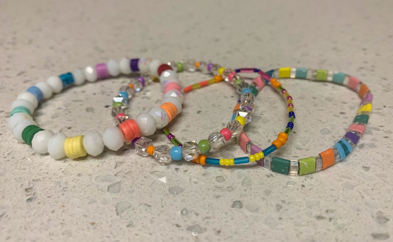 May include: Four colorful beaded bracelets, one with white beads, one with clear crystal beads, one with small square beads in a rainbow pattern, and one with a mix of blue, green, and orange beads.
