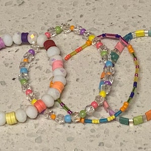 May include: Four colorful beaded bracelets with white beads and various colored beads. The bracelets are stacked on top of each other.