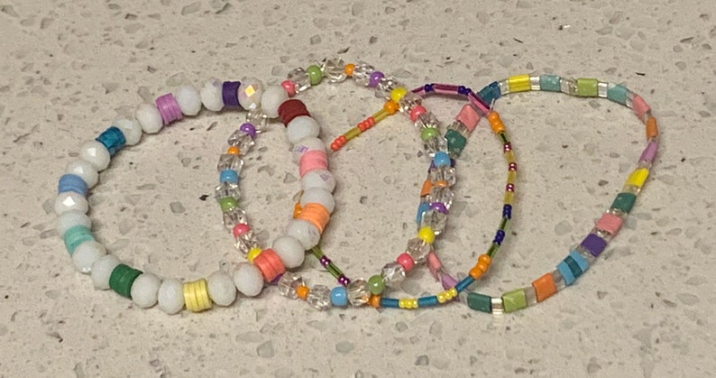 May include: Four colorful beaded bracelets with white beads and various colored beads. The bracelets are stacked on top of each other.