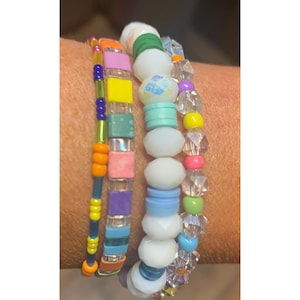 May include: A set of four colorful beaded bracelets. The bracelets feature a variety of shapes and sizes of beads, including square, round, and oval beads. The beads are in shades of pink, blue, green, yellow, orange, and purple.