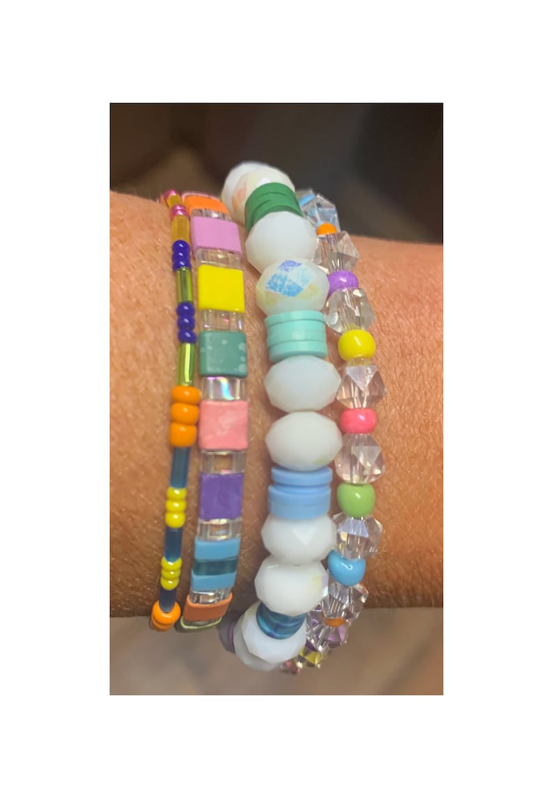 May include: A set of four colorful beaded bracelets. The bracelets feature a variety of shapes and sizes of beads, including square, round, and oval beads. The beads are in shades of pink, blue, green, yellow, orange, and purple.