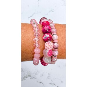 Op de afbeelding: Vier roze en rode kralenarmbanden gestapeld op een pols. De armbanden zijn gemaakt van verschillende materialen, waaronder glas, agaat en plastic.