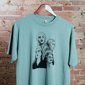 May include: A light blue t-shirt with a black and white illustration of four women's faces. The women are looking in different directions and have different hairstyles. The t-shirt is hanging on a hanger against a brick wall.