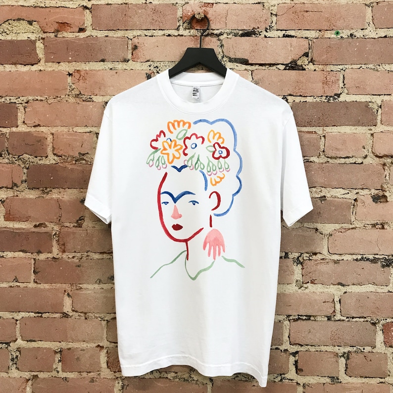 May include: White t-shirt with a colorful illustration of Frida Kahlo wearing a flower crown. The illustration is in a simple, modern style.
