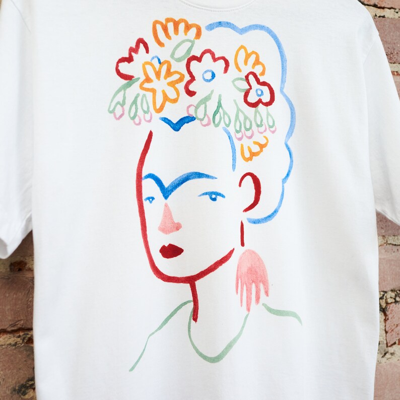 May include: White t-shirt with a colorful illustration of a woman with a flower crown. The woman has blue eyes, red lips, and a red eyebrow. The flowers are in shades of red, orange, yellow, blue, and green.