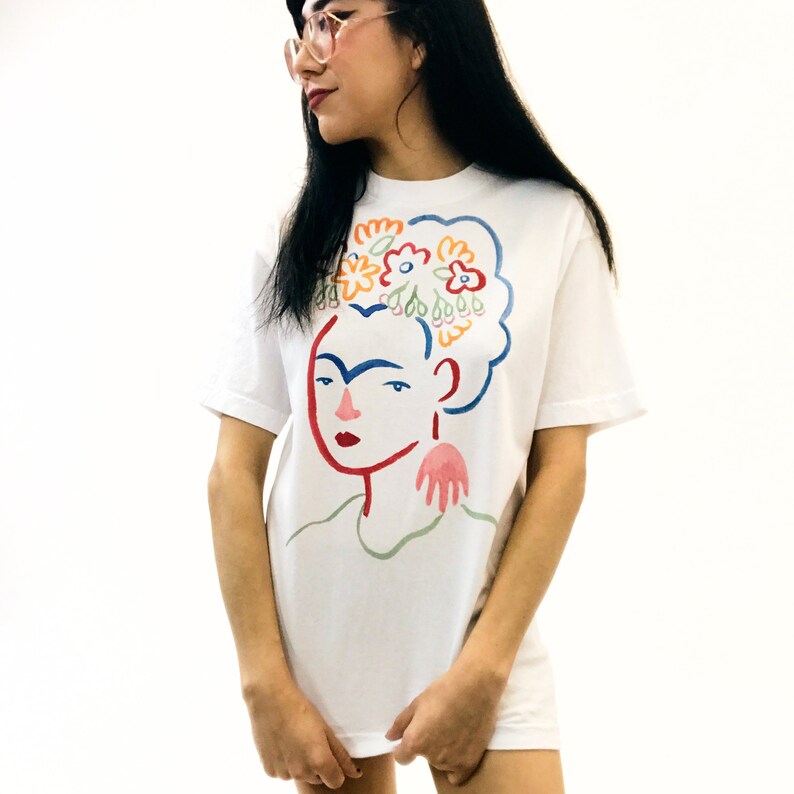 May include: White t-shirt with a colorful illustration of Frida Kahlo with flowers in her hair.
