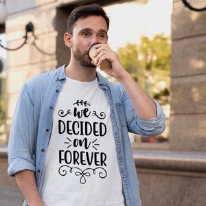 Könnte beinhalten: Weißes T-Shirt mit dem Text "We Decided on Forever" in schwarzer, dekorativer Schrift. Das Shirt wird von einer Person getragen, die ein hellblaues Jeanshemd trägt. Die Person hält einen braunen Kaffeebecher aus Papier.
