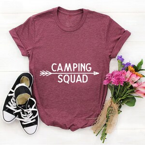 May include: A maroon t-shirt with the text "CAMPING SQUAD" in white lettering with an arrow pointing to the right and left. The shirt is laid flat on a white surface with a bouquet of flowers and two black trainers.