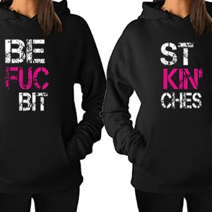 May include: Two black hooded sweatshirts with a matching design. The left sweatshirt says "BE FUC BIT" in white and pink lettering. The right sweatshirt says "ST KIN CHES" in white and pink lettering.