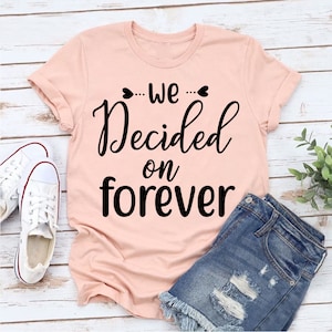 Könnte beinhalten: Ein rosa T-Shirt mit dem Text "...we... Decided on forever" in schwarzer Schrift.