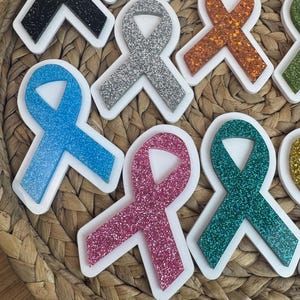 May include: A collection of colorful awareness ribbon-shaped ornaments with glittery surfaces. The ribbons are in various colors, including blue, pink, silver, and green, each set within a white border. The ornaments are arranged on a woven, textured surface.