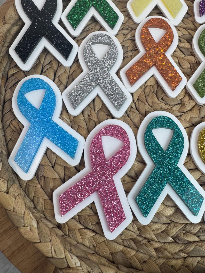 May include: A collection of colorful awareness ribbon-shaped ornaments with glittery surfaces. The ribbons are in various colors, including blue, pink, silver, and green, each set within a white border. The ornaments are arranged on a woven, textured surface.
