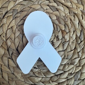 May include: A white ribbon-shaped pin with a circular emblem featuring a bee design. The pin is set against a woven, textured background, creating a contrast in textures and colors. The pin is likely a symbol of awareness or support.
